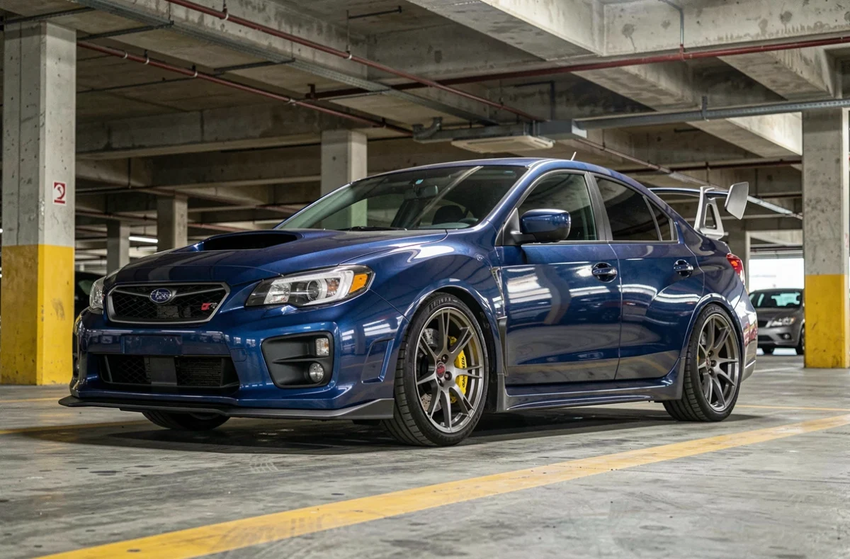 Subaru Impreza STI bleu foncé dans un parking urbain en béton, vue grand angle au niveau du pare-chocs révélant l'aileron arrière iconique et les étriers Brembo dorés